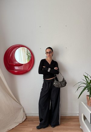 Svart långärmad zip-up tröja, lösa svarta byxor med vita ränder, liten grå handväska, rund röd spegel, inomhusväxt i kruka.