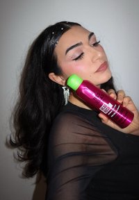 Pink spray bottle with green cap labeled "Bad Head Rush," held against a black sheer top. Hair cascading with scattered decorative beads.