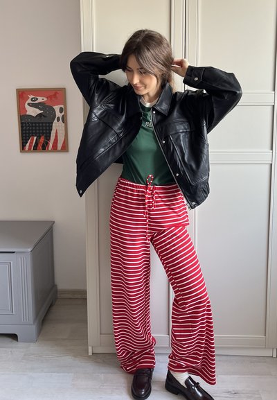 Chaqueta oversized de cuero negro, camisa verde, pantalones de pierna ancha a rayas rojas y blancas, y zapatos oscuros con tacón bajo. Fondo neutro.