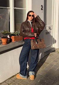 Femme en veste en cuir marron foncé et lunettes de soleil debout devant une porte blanche, tenant un sac à main imprimé léopard dans une rue pavée.