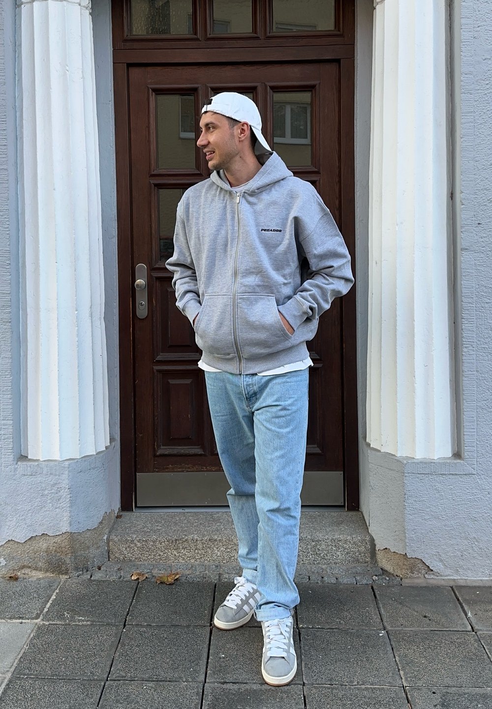 Grå zip-up hettegenser med svart logo, iført over en hvit skjorte, kombinert med lyseblå jeans og grå sneakers. Står foran en trekledd dør.