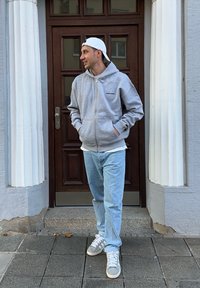 Grå zip-up hettegenser med svart logo, iført over en hvit skjorte, kombinert med lyseblå jeans og grå sneakers. Står foran en trekledd dør.