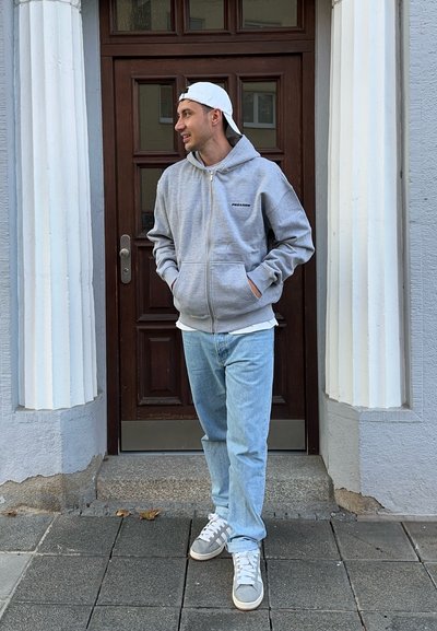 Sudadera gris con cremallera y logo negro, llevada sobre una camisa blanca, combinada con jeans azul claro y zapatillas grises. De pie frente a una puerta de madera.