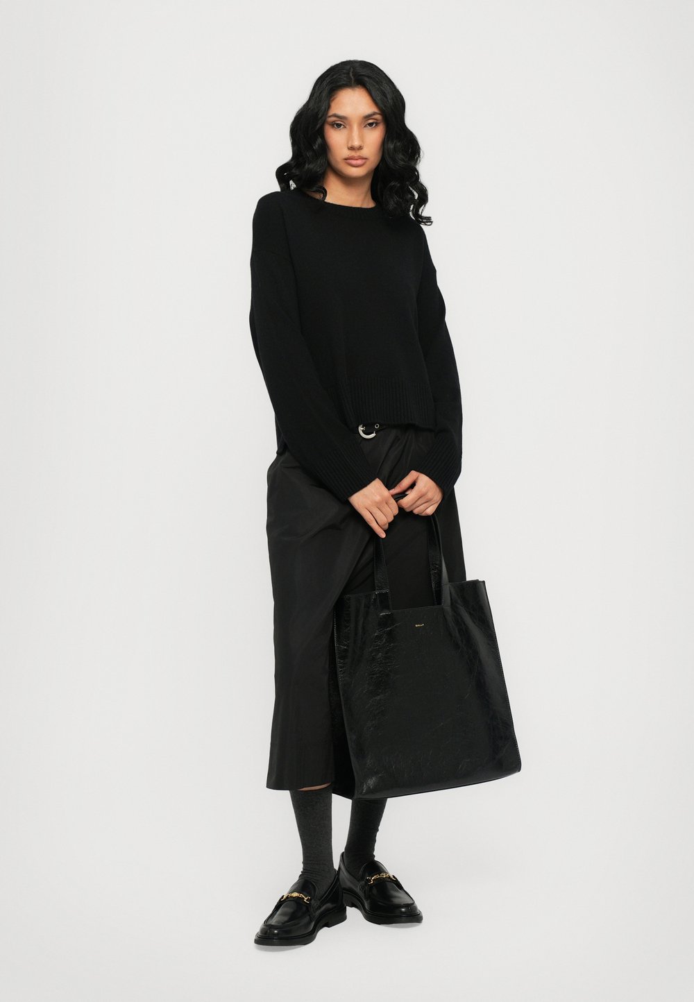 Maglione nero lavorato a maglia con maniche lunghe, abbinato a pantaloni neri larghi e una borsa a tracolla nera lucida. Il modello indossa mocassini neri e calzini grigi.