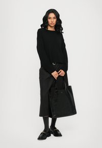Maglione nero lavorato a maglia con maniche lunghe, abbinato a pantaloni neri larghi e una borsa a tracolla nera lucida. Il modello indossa mocassini neri e calzini grigi.