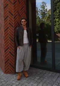 Brun lærjakke, hvit tanktop, grå joggebukser og brune støvler. Står mot en murvegg med glassrefleksjon.