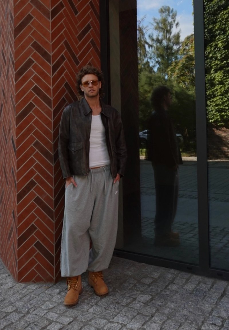 Braune Lederjacke, weisses Tanktop, graue Jogginghose und hellbraune Stiefel. Steht gegen eine Backsteinmauer mit einer Glasreflexion.