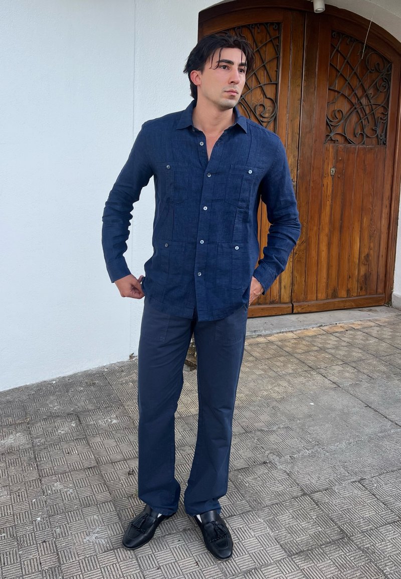 Homme debout à l'extérieur près d'une porte en bois, portant une chemise bleu foncé à boutons, un pantalon foncé assorti et des mocassins noirs.
