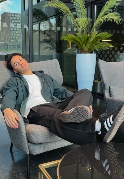 Sillón de tela gris con una textura suave, pantalones negros, sudadera verde con cremallera, y zapatos negros con rayas blancas. Mesa de cristal y planta de palma visibles.