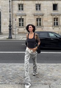 Top nero senza maniche con spalline borchiate, abbinato a pantaloni bianchi con un motivo ondulato grigio. Pavimentazione in cemento e sfondo urbano.