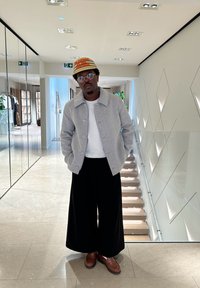 Άτομο ντυμένο με ένα ανοιχτό γκρι μάλλινο μπουφάν, λευκή μπλούζα και μαύρο παντελόνι με φαρδιά γραμμή, με καφέ παπούτσια και καπέλο με ρίγες. Σύγχρονη εσωτερική διακόσμηση.