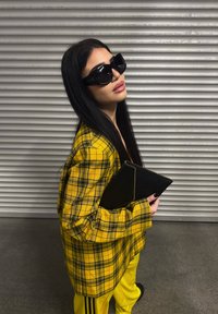 Yellow check oversized shirt, black zippered clutch, black sunglasses, long straight black hair, and yellow jogging bottoms.