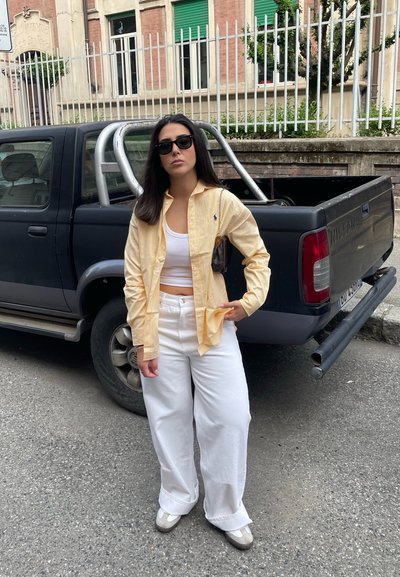 Camisa de botones a rayas crema y amarilla sobre una camiseta blanca sin mangas, combinada con pantalones anchos blancos y zapatos de color claro. Gafas de sol negras puestas.