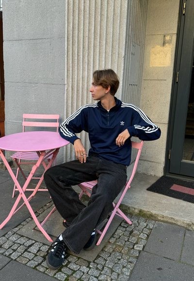 Sudadera azul marino con media cremallera y rayas blancas, jeans negros de pierna ancha y zapatillas negras, sentado en una mesa plegable rosa en un ambiente urbano.