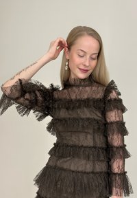 Juoda nėrinių palaidinė su aukštu kaklu ir raukiniais rankovėse, dengtose sluoksniuotais raukiniais. Permatoma medžiaga su sudėtingais gėlių raštais ir tekstūra.