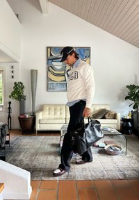 Mann in weißem Sweatshirt und schwarzer Mütze, der im Wohnzimmer geht und eine schwarze Lederhandtasche trägt, mit beiger Couch und abstrakter Malerei im Hintergrund.