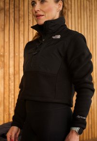 Svart genser med fleece og glidelås, strukturert stoff og logo-detaljer. Kombinert med tettsittende svarte bukser som fremhever et sporty utseende.