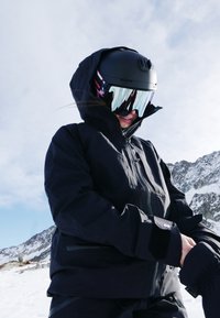 Schwarze Skijacke mit hohem Kragen, Reißverschluss und elastischen Bündchen, kombiniert mit einem schwarzen Helm und reflektierenden Skibrillen, auf schneebedecktem Gelände.