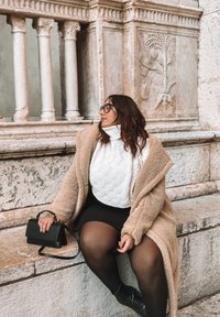 Casaco de shearling bege sobre um suéter branco de ponto de cabos, saia mini preta e meias pretas. Asegurando uma pequena mala preta, sentada em degraus de pedra.