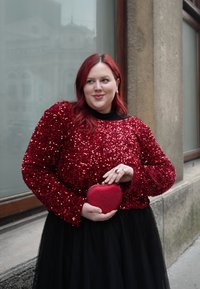 Femme aux cheveux roux portant un haut rouge à paillettes et une jupe noire, tenant un sac rouge en forme de cœur, debout près d'une fenêtre de bâtiment.