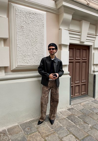 Persona vistiendo una chaqueta de cuero negra, pantalones de estampado de leopardo y zapatos de punta negros, de pie contra una pared texturizada de color rosa claro.
