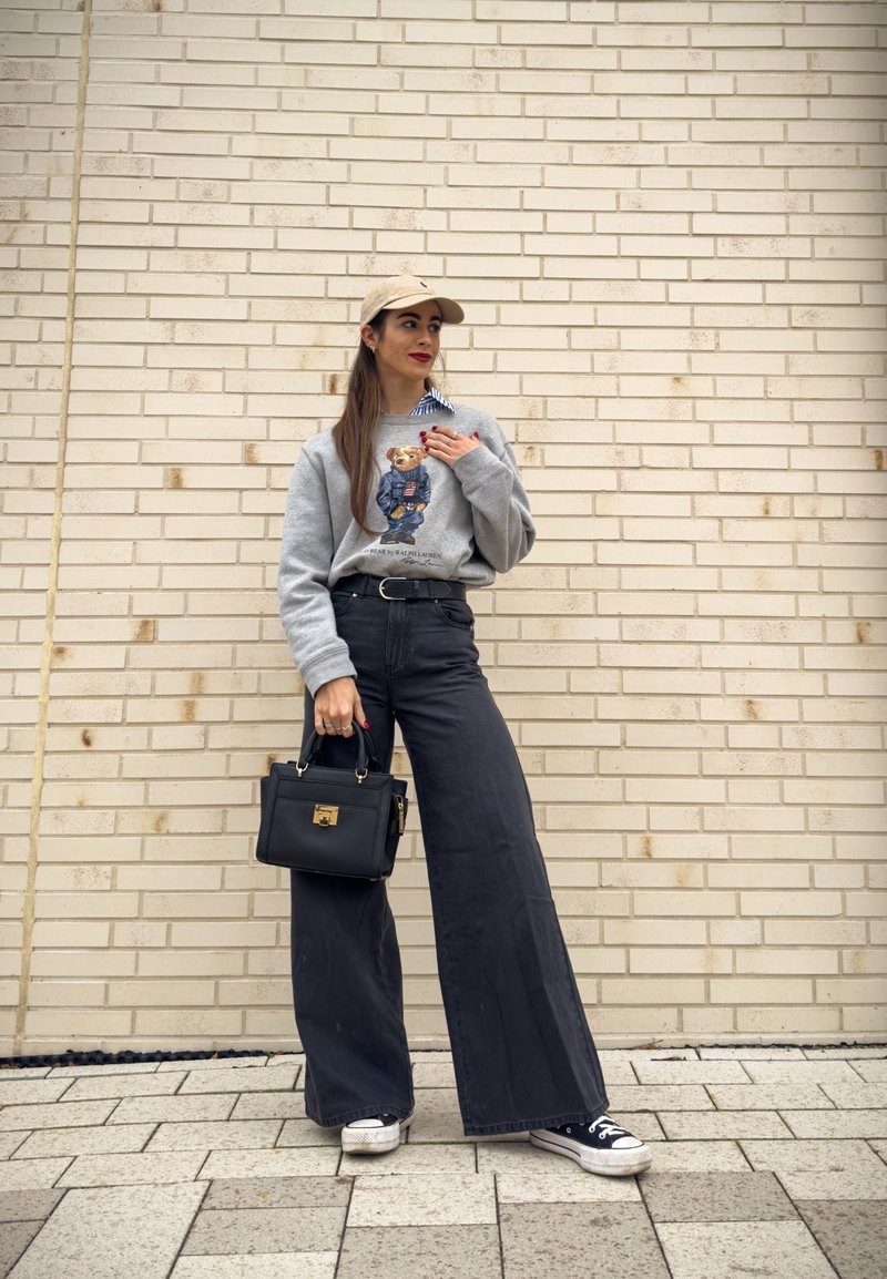 Grå sweatshirt med en björngrafik, svart vida byxor med hög midja, svart handväska med gulddetaljer och svarta sneakers.