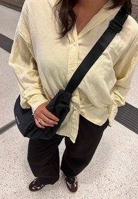 Sac bandoulière noir avec sangle réglable, chemise jaune texturée à boutons, pantalon large marron foncé, et chaussures à motifs.