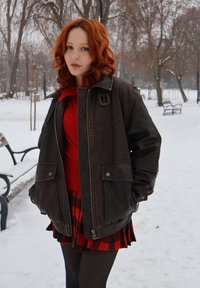 Jeune femme aux cheveux roux bouclés, portant une veste en cuir marron, un haut rouge et une jupe à carreaux, debout dans un parc enneigé avec des arbres dénudés et des bancs.