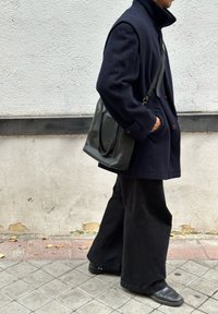 Abrigo de lana azul marino con cuello alto, pantalones anchos negros y bolso de cuero negro, caminando sobre una acera de baldosas frente a una pared texturizada.