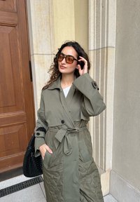 Πράσινο καπιτονέ παλτό με ζωνάκι στη μέση, μεγάλα κουμπιά και φαρδιά μανίκια. Συνδυασμένο με υπερμεγέθεις καφέ γυαλιά ηλίου και μια μαύρη πλεκτή τσάντα.