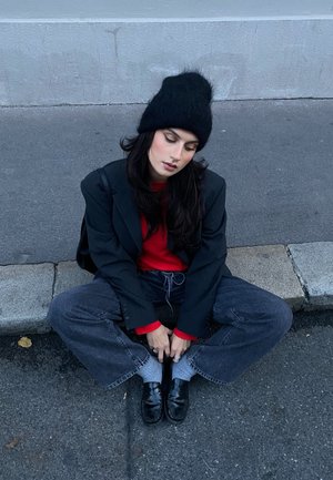 Sombrero de lana negro, camiseta roja y blazer negro. Jeans oscuros sueltos, calcetines grises y zapatos negros. Sentado en el pavimento con las piernas cruzadas.