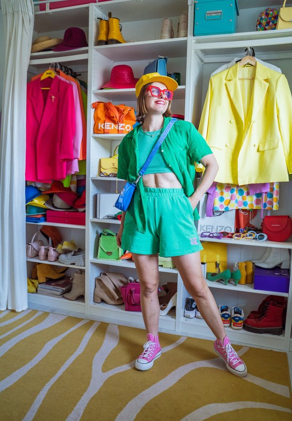 Kvinde i grønt outfit, gul kasket og pink sko står smilende i en farverig walk-in closet med tøj, sko og accessories på hylderne.