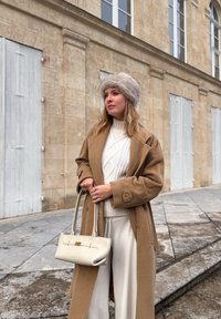 Γυναίκα ντυμένη με καφέ παλτό, λευκό πλεκτό πουλόβερ, λευκό παντελόνι και γκρίζο γούνινο καπέλο, κρατώντας ένα λευκό τσαντάκι, στέκεται σε πέτρινο πεζοδρόμιο κοντά σε παλιό κτίριο.