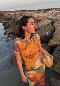 Abito aderente con un vivace motivo floreale in tonalità di arancione, giallo e verde. Tessuto morbido; la modella tiene una piccola borsa beige. Sfondo di una costa rocciosa.