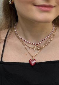 Collana con pendente a forma di cuore rosso, con dettagli in strass, stratificata con collane di perle e catene d'oro, caratterizzata da varie texture e dimensioni.