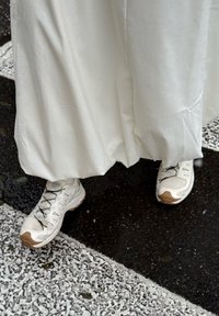 Pantaloni bianchi ampi che coprono scarpe da ginnastica grigio chiaro con lacci neri e dettagli color cuoio, su uno sfondo scuro e strutturato.