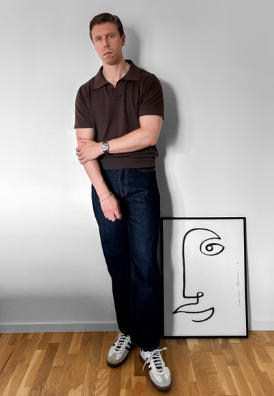 Hombre con camisa marrón oscuro y vaqueros, de pie con los brazos cruzados junto a un dibujo abstracto de rostro minimalista enmarcado en blanco y negro sobre suelo de madera.