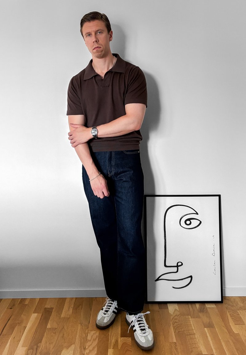 Homme en chemise marron foncé et jeans, les bras croisés, debout à côté d'un dessin abstrait minimaliste de visage en noir et blanc encadré sur un sol en bois.