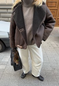 Καφέ τζάκετ με επένδυση από μαλλί και κολάρο, φορεμένο πάνω από φωτεινό καφέ ριγωτό πουλόβερ, συνδυασμένο με λευκά παντελόνια από βελούδο και μαύρες μποτάκια μέχρι τον αστράγαλο. Περιλαμβάνεται και μαύρη τσάντα.