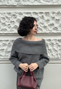 Kvinna med lockigt mörkt hår i en grå av-axlarna tröja som håller en liten vinröd handväska mot en texturerad ljus vägg.
