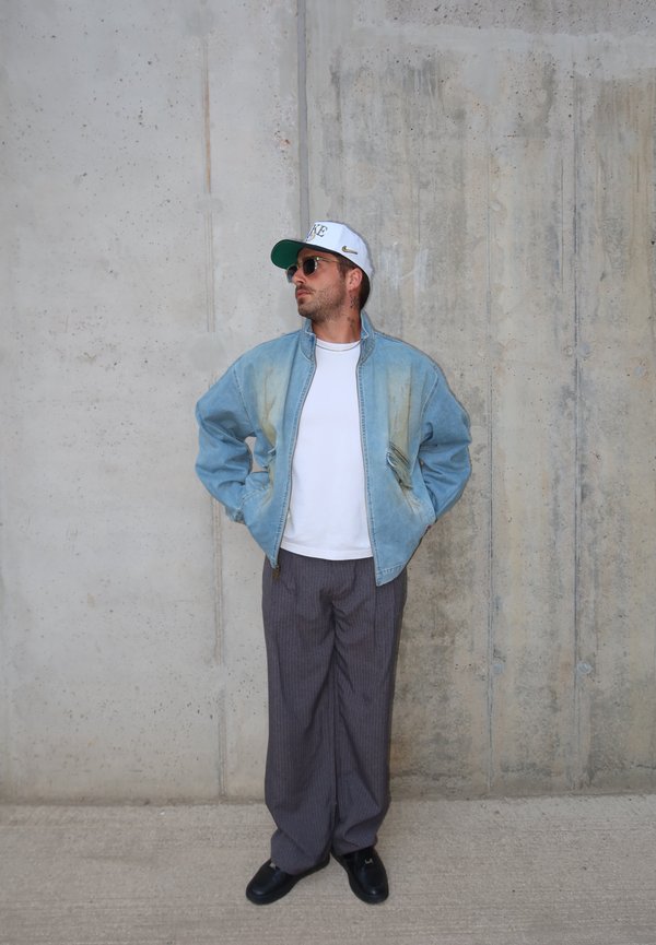 Uomo con cappello bianco, occhiali da sole, giacca azzurra, camicia bianca, pantaloni grigi e scarpe nere, in piedi contro un muro di cemento con le mani in tasca.