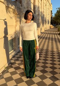 Femme aux cheveux noirs coupés en carré, portant un haut blanc à manches longues et un pantalon de survêtement Adidas vert, tenant un petit sac à main rose sur un trottoir à motif.