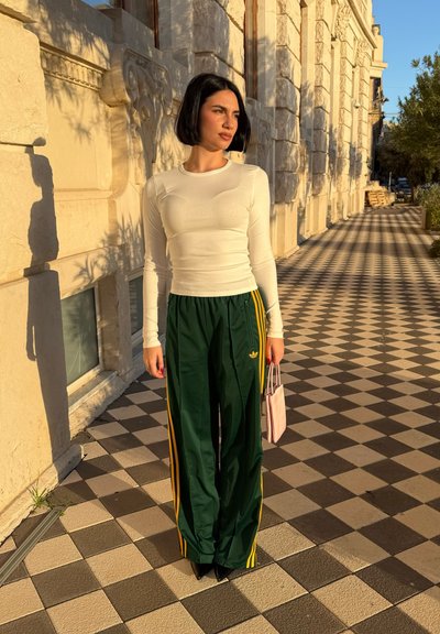 Mujer con cabello corto negro, vestida con una blusa blanca de manga larga y pantalones deportivos verdes de Adidas, sosteniendo un pequeño bolso rosa en una acera con diseño.