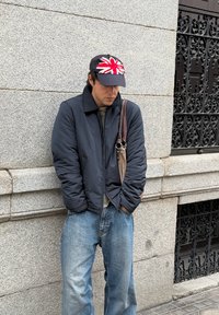 Veste noire matelassée avec col, chemise grise en dessous, jean bleu ample, et casquette noire avec un motif Union Jack rouge et blanc.
