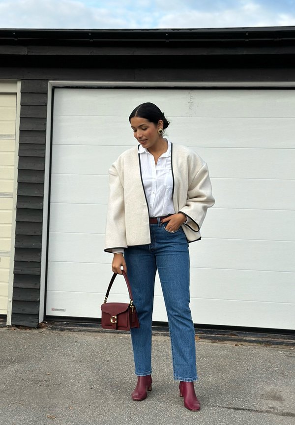 Lichtgrijze oversized jas met zwarte randen, witte blouse, blauwe jeans, bordeauxrode handtas en bordeauxkleurige enkellaarsjes op een persoon die buiten staat.