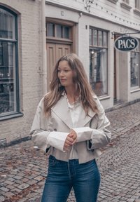 Heleroosa lühike trençkot suurte kraedega, topeltkangaste ja laiade varrukatega, kantud valge särke ja siniste teksade peal. Munakivitee.