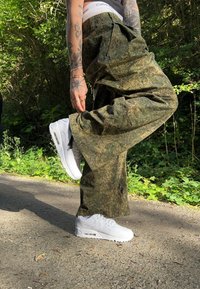 Kamufliažo krovinių kelnės su laisvu siluetu žemės atspalviuose, derinamos su baltais Nike Air Max batais, turinčiais matomą oro pagalvę pado.