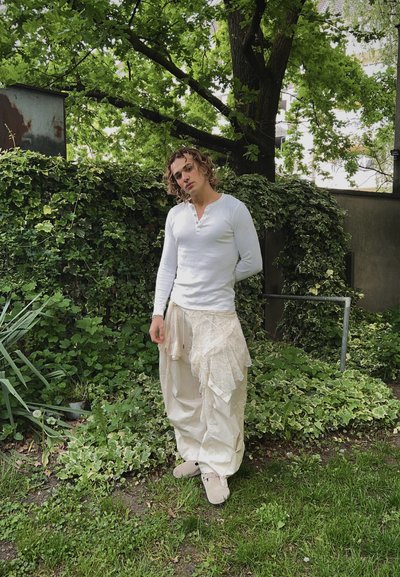 Camisa henley de manga larga blanca, pantalones cremosos texturizados con detalles en capas y calzado ligero, situados ante un frondoso fondo de vegetación verde.