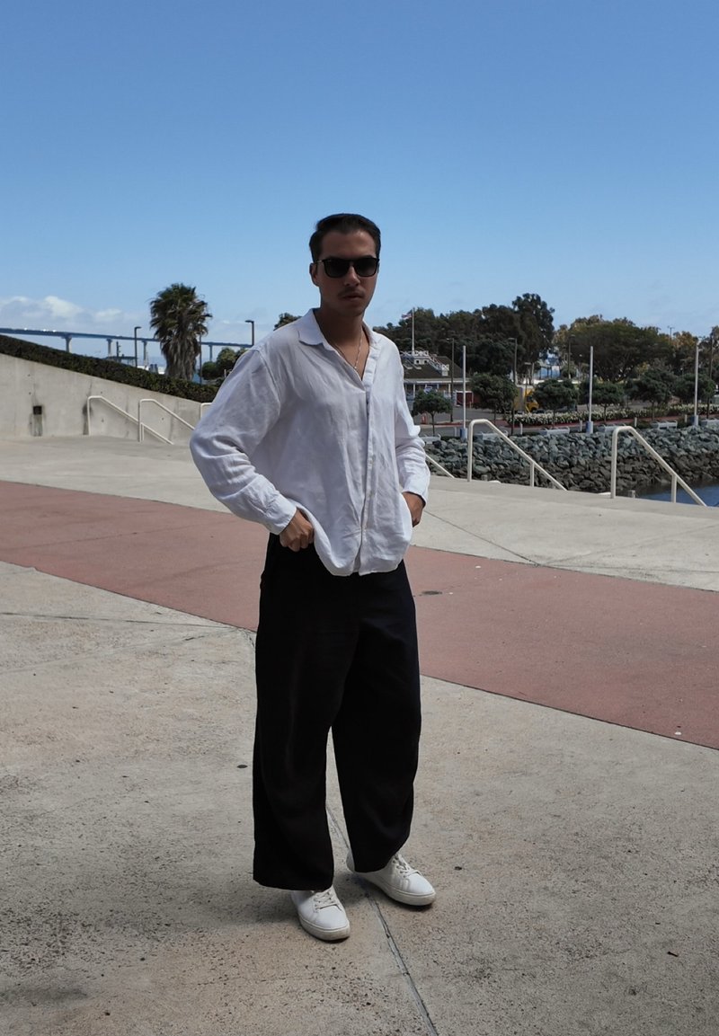 Homme portant des lunettes de soleil, une chemise blanche, un pantalon large noir et des chaussures blanches, debout sur une surface pavée près de l'eau et des arbres sous un ciel clair.