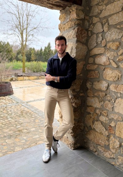 Joven con una sudadera con cremallera azul marino y pantalones beiges, apoyado contra una pared de piedra. Lleva zapatillas blancas y tiene un reloj en la muñeca.